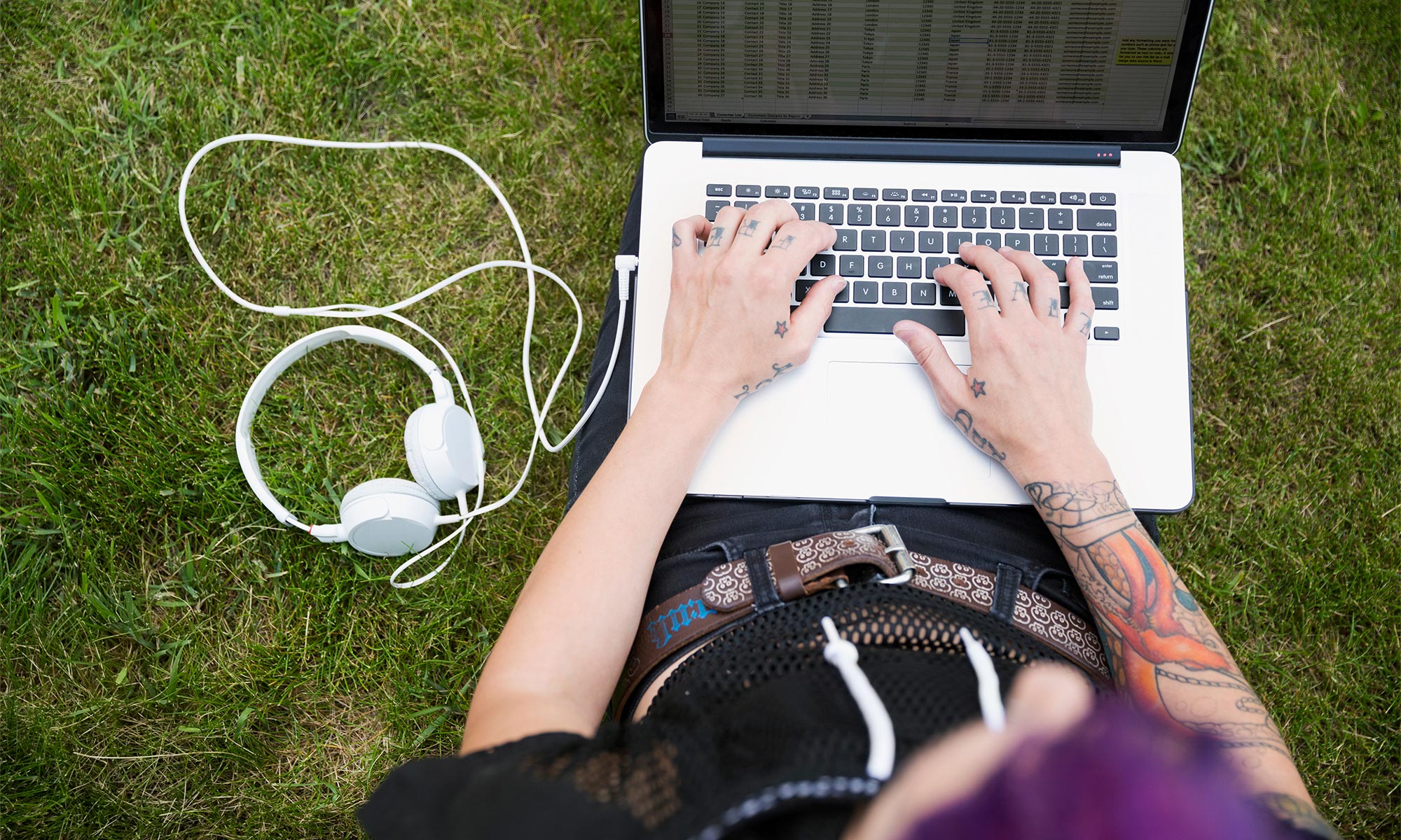 Ung person med tatueringar sitter ute i gräset med en laptop i knät